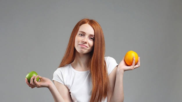 Retrato de una joven de pie sobre un fondo gris se preguntó qué fruta es más saludable Apple o naranja