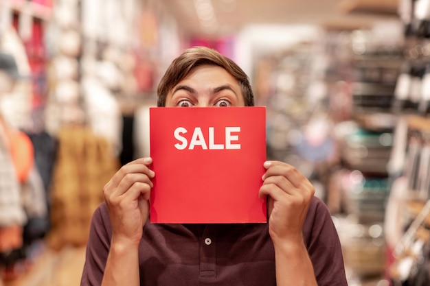 Foto retrato de un joven posa con un cartel de venta rojo, cubriéndose la cara con él, en la sala de comercio. venta. descuentos descuentos locos. emociones de deleite.