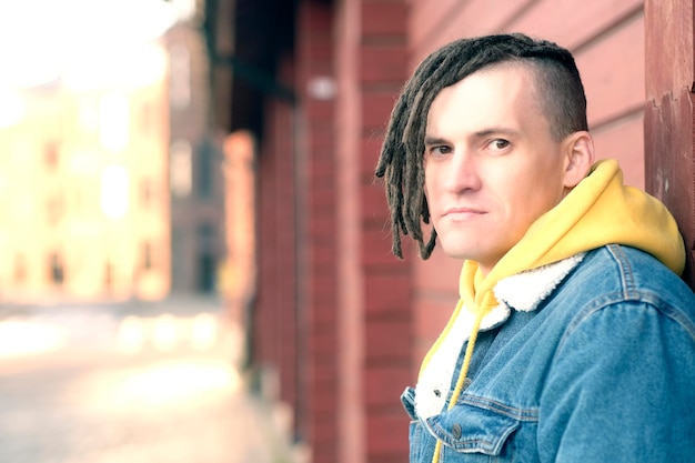 Retrato de joven positivo con rastas de pie apoyado en la pared en la calle de la ciudad