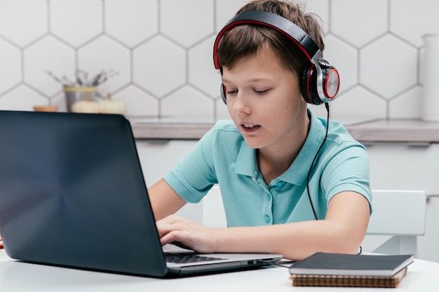 Retrato de un joven preadolescente con audífonos sentado en el escritorio cerca de cuadernos usando una computadora portátil con una lección en línea