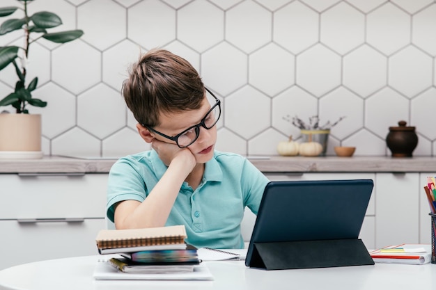 Retrato de un joven preadolescente concentrado sentado en el escritorio cerca del libro de lectura del bloc de notas cerca de la tableta que tiene una lección en línea