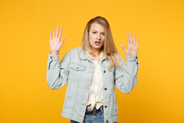 Retrato de una joven preocupada con ropa casual de denim mirando una cámara, levantando las manos, mostrando las palmas aisladas en un fondo naranja amarillo brillante en el estudio. Concepto de estilo de vida de las personas. Simulacros de espacio de copia.