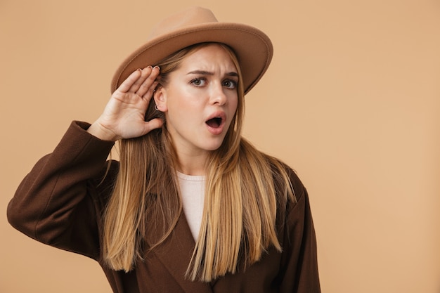 Retrato de joven preocupada con sombrero y abrigo preguntándose mientras escucha con su mano aislada en beige