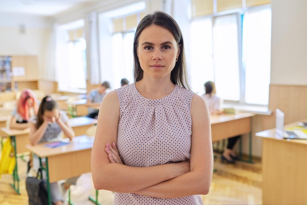 Retrato de una joven profesora sonriente mirando la cámara con los brazos cruzados en el aula con los estudiantes. Educación, escuela, universidad, concepto de enseñanza.