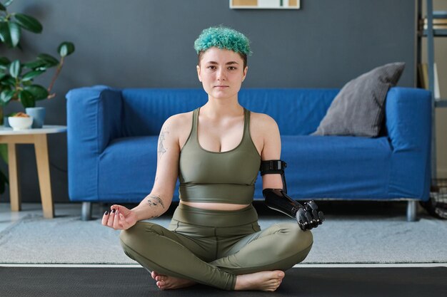 Retrato de una joven con prótesis sentada en una alfombra de ejercicios en la habitación y haciendo yoga
