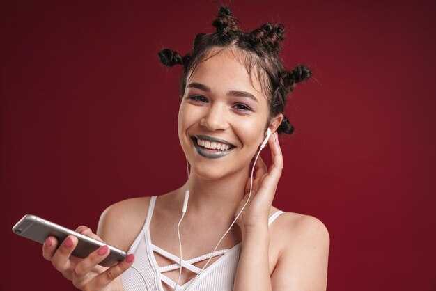 Retrato de joven punk con peinado extraño y lápiz labial oscuro riendo mientras usa un teléfono inteligente con auriculares aislados sobre una pared roja