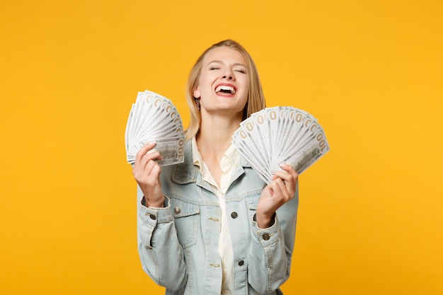 Retrato de una joven que se ríe con ropa informal de denim sostiene un abanico de dinero en efectivo en billetes en dólares aislados en un fondo de pared naranja amarillo en el estudio. Concepto de estilo de vida de las personas. Simulacros de espacio de copia.