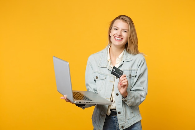 Retrato de una joven que se ríe con ropa informal de mezclilla sosteniendo una computadora portátil, tarjeta de crédito aislada en un fondo de pared naranja amarillo en el estudio. Concepto de estilo de vida de las personas. Simulacros de espacio de copia.