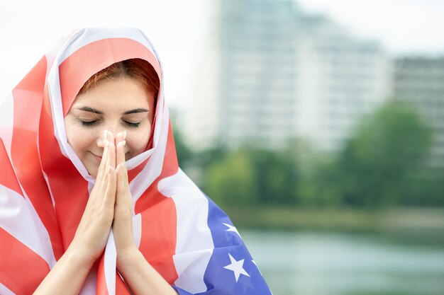 Retrato de joven refugiada con la bandera nacional de Estados Unidos en la cabeza y los hombros. Chica musulmana positiva rezando al aire libre.