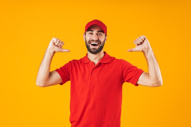 Retrato de joven repartidor en camiseta roja y gorra sonriendo y señalando con el dedo a sí mismo aislado sobre amarillo