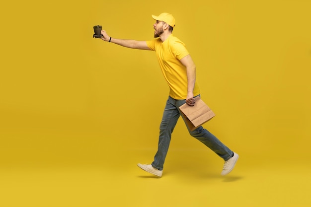 Retrato de joven repartidor en uniforme amarillo con paquete de papel y café para llevar aislado sobre pared amarilla