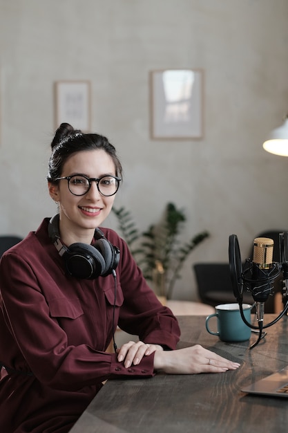 Retrato de joven reportera sentada en su lugar de trabajo frente al micrófono y sonriendo a la cámara durante su trabajo en el estudio de radio