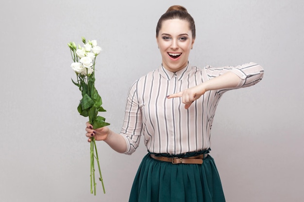 Retrato de una joven romántica sorprendida y atractiva con camisa a rayas y falda verde sosteniendo un ramo de flores blancas y señalando con el dedo. Interior, foto de estudio, aislado sobre fondo gris, espacio de copia