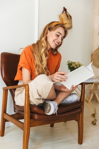 Retrato de una joven rubia alegre en casa en el interior leyendo una revista sentado en una silla.