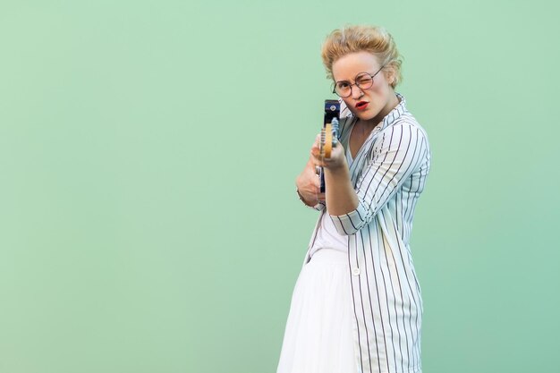 Retrato de una joven rubia atenta y divertida con blusa blanca a rayas con anteojos de pie, sosteniendo guitarra eléctrica, mirando y centrándose en la cámara. tiro del estudio aislado sobre fondo verde claro