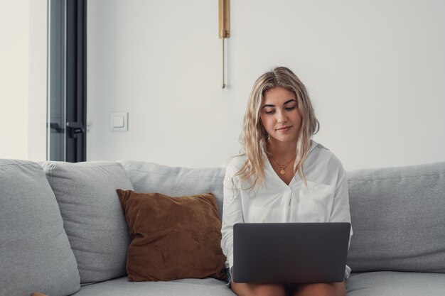 Retrato de una joven rubia atractiva usando una computadora portátil en el sofá relajándose navegando por el