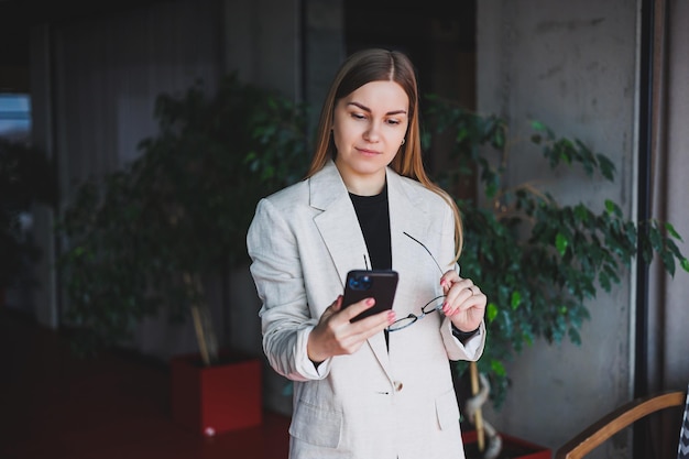 Retrato de una joven rubia con una chaqueta beige y gafas en la cara un teléfono mod en manos de una mujer Hermosa chica bloguera moderna