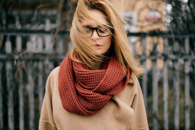 Retrato de joven rubia elegante con gafas y bufanda posando al aire libre