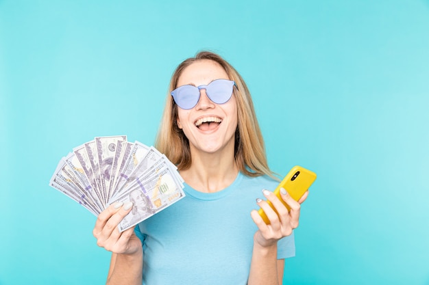 Retrato de una joven rubia emocionada mostrando un montón de billetes de banco y sosteniendo un teléfono móvil aislado en azul