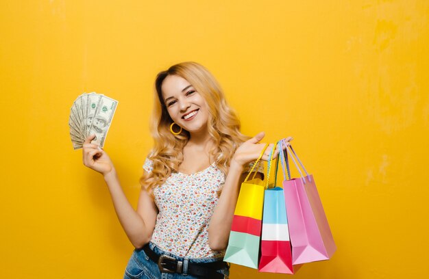 Retrato de una joven rubia feliz celebración de billetes y bolsa de compras sobre pared amarilla