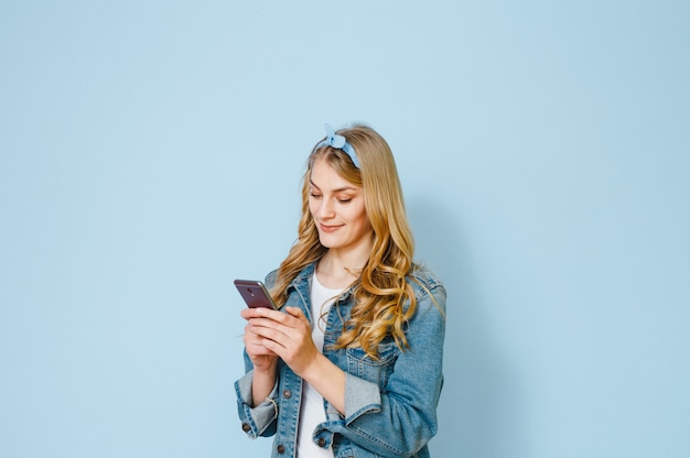 Retrato de una joven rubia feliz por qué ve en su teléfono móvil aislado sobre fondo azul.