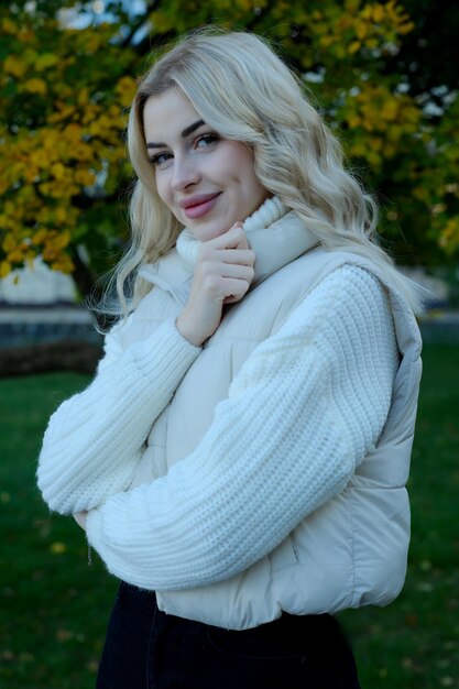 Retrato de una joven rubia hermosa con cabello largo en un suéter blanco y chaqueta en un otoño soleado en un parque de la ciudad