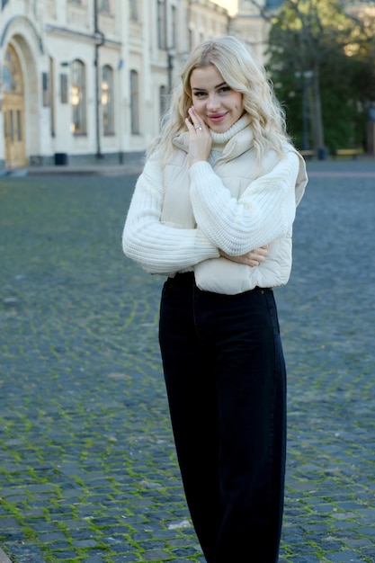 Retrato de una joven rubia hermosa con cabello largo en un suéter blanco y chaqueta en un otoño soleado en un parque de la ciudad