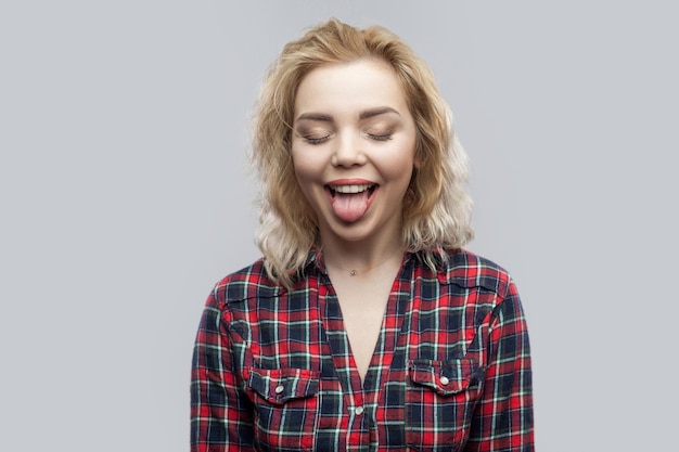 Retrato de una joven rubia hermosa y graciosa con un pantalones a cuadros rojo casual de pie con los ojos cerrados y la lengua afuera. tiro de estudio interior, aislado sobre fondo gris.