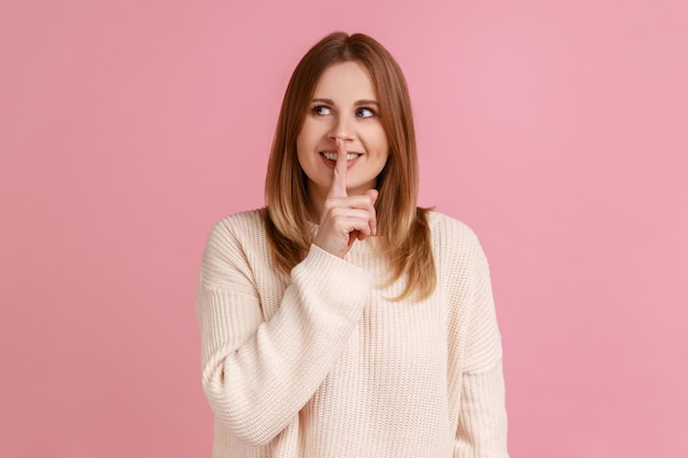 Retrato de una joven rubia optimista y positiva de pie mostrando un signo de silencio y mirando hacia otro lado compartiendo un secreto usando un suéter blanco Foto de estudio interior aislada en un fondo rosa