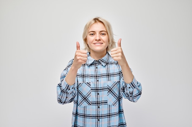 Retrato de una joven rubia con ropa informal sonriendo alegremente mostrando los pulgares hacia arriba gesto aislado en el fondo blanco del estudio Aprueba una buena elección decisión correcta