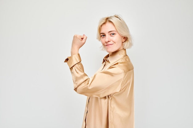 Retrato de una joven rubia segura de sí misma mostrando bíceps demostrando fuerza aislada en el fondo blanco del estudio Concepto de ganador afortunado