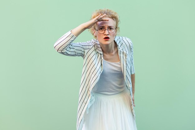 Retrato de una joven rubia seria con camisa blanca, falda y blusa a rayas con anteojos de pie con la mano en la frente y mirando atenta. tiro del estudio aislado sobre fondo verde claro.