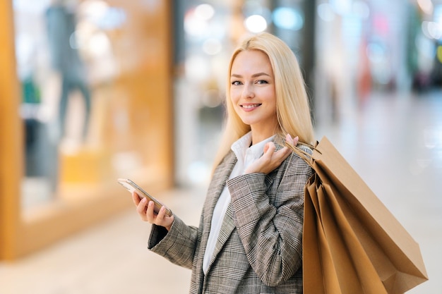 Retrato de una joven rubia sonriente con un elegante abrigo mirando a la cámara sosteniendo un teléfono móvil y bolsas de papel de compras con compras en el salón del centro comercial de fondo borroso