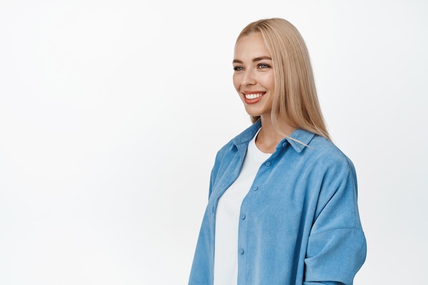 Retrato de una joven rubia con una sonrisa blanca mirando a un lado con una expresión feliz y relajada en la cara, una pose natural casual de pie sobre un fondo blanco