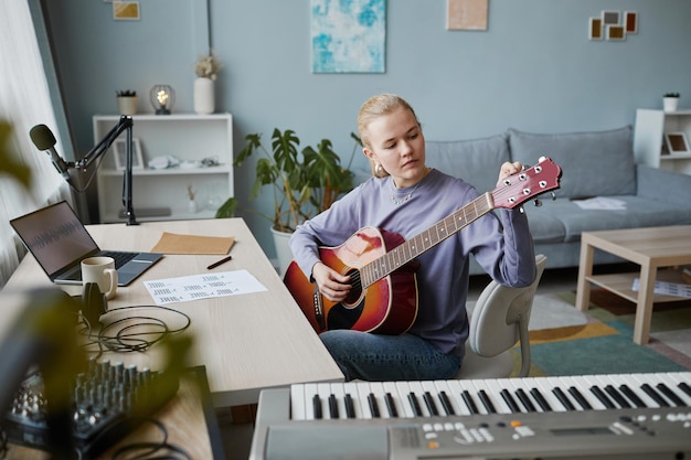 Retrato de una joven rubia tocando la guitarra en casa y componiendo una pista de música usando una grabación suave