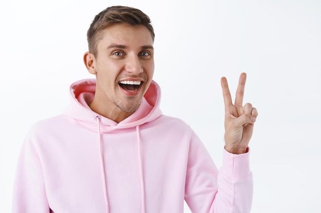 Retrato de un joven rubio guapo, vestido con una sudadera con capucha rosa, que se mantiene en el lado positivo, que es positivo, que muestra el signo de buena voluntad de la paz y que sonríe ampliamente, que disfruta de una fiesta increíble, que se coloca en la pared blanca