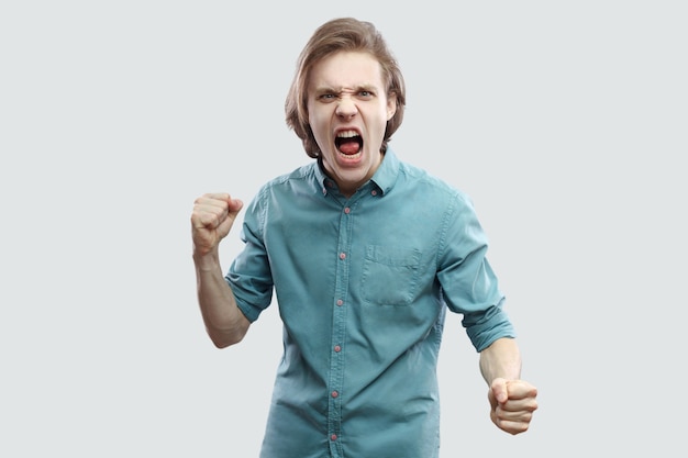 Retrato de joven rubio de pelo largo guapo loco enojado en camisa casual azul de pie, mirando a cámara, gritando y listo para atacar. tiro del estudio de interior, aislado en fondo gris claro.