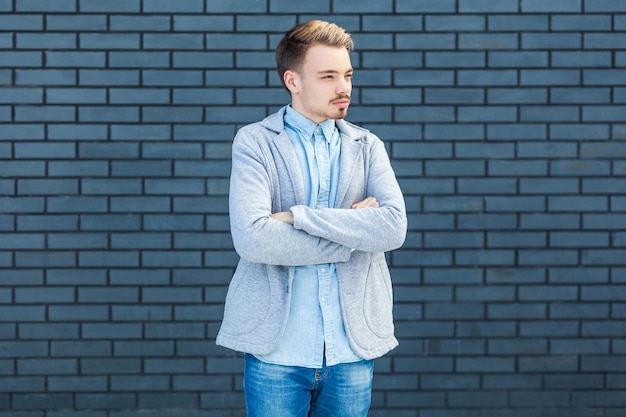 Retrato de un joven rubio, tranquilo y serio, de estilo informal, de pie con los brazos cruzados y mirando hacia otro lado con la cara relajada. foto de estudio interior sobre fondo de pared de ladrillo.