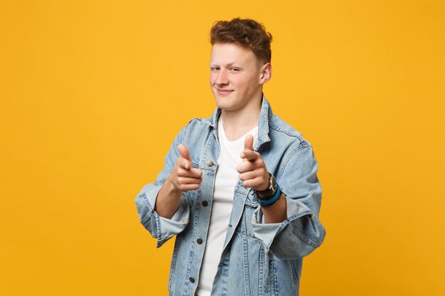 Retrato de un joven seguro de sí mismo con ropa informal de denim que señala con el dedo índice a la cámara aislada en un fondo amarillo anaranjado en el estudio. Emociones sinceras de la gente, concepto de estilo de vida. Simulacros de espacio de copia.