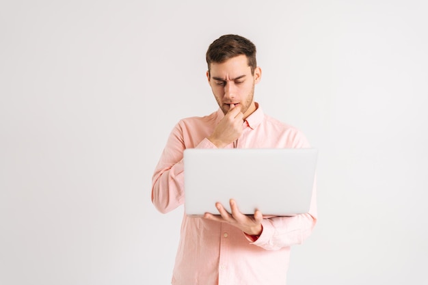 Retrato de un joven serio y reflexivo de pie con una laptop mirando en la pantalla con una cara confundida y desconcertada sobre un fondo blanco aislado. Disparo de estudio de hombre pensativo posando con pc mirando la pantalla