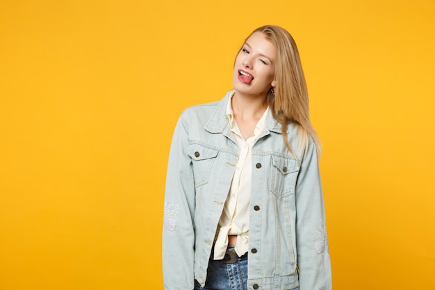 Retrato de una joven sexy y divertida con ropa informal de denim mirando la cámara, mostrando la lengua, parpadeando aislada en un fondo naranja amarillo brillante en el estudio. Concepto de estilo de vida de las personas. Simulacros de espacio de copia.