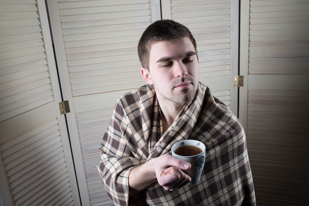 Retrato de un joven somnoliento envuelto en una tela escocesa sosteniendo una taza de té en sus manos sobre la superficie del armario