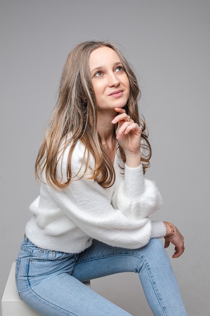 Retrato de joven soñadora caucásica de pelo largo en suéter blanco y jeans en la pared gris