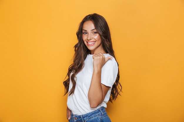 Retrato de una joven sonriente con cabello largo morena de pie sobre la pared amarilla, apuntando hacia afuera