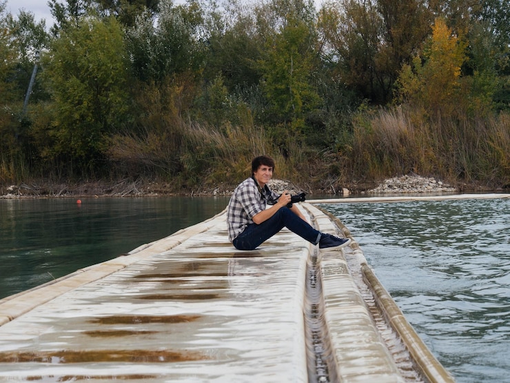 Foto retrato de un joven sonriente con una cámara sentado en el camino en medio del lago