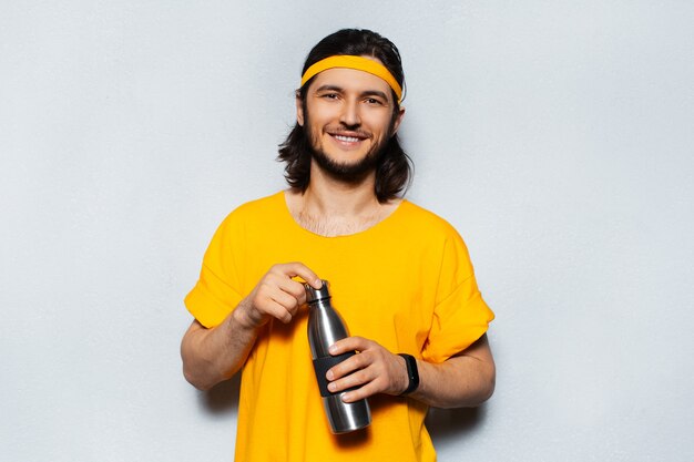 Retrato de joven sonriente con camiseta naranja sosteniendo una botella de aluminio sobre fondo blanco.