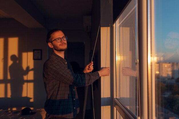 Retrato de un joven sonriente cierra persianas enrolladas en la ventana en un día soleado al atardecer