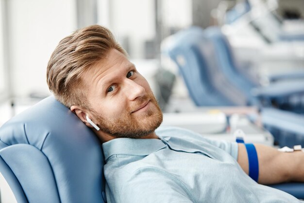 Retrato de un joven sonriente donando sangre mientras está acostado en una silla cómoda en el centro médico y mira