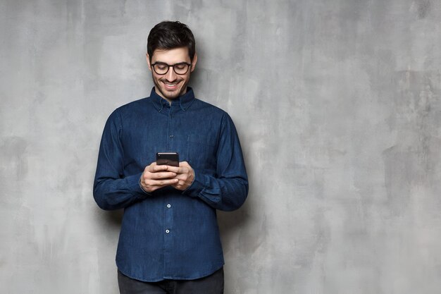 Foto retrato de un joven sonriente escribiendo un mensaje con una nueva aplicación en su teléfono inteligente de pie contra una pared de textura gris con espacio para copiar