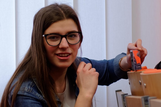 Foto retrato de una joven sonriente fotografiando a través de un teléfono inteligente contra persianas